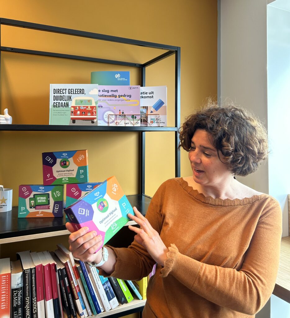 Persoon staat voor een boekenkast met Optimaal Digitaal spellen en heeft in haar handen de nieuwste versie van het spel vast